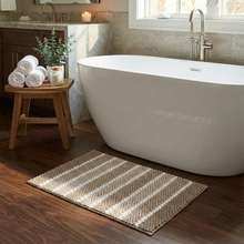تحميل الصورة في عارض المعرض، Bathroom with freestanding bathtub, wooden stool with towels and plant, and striped rug on wooden floor.
