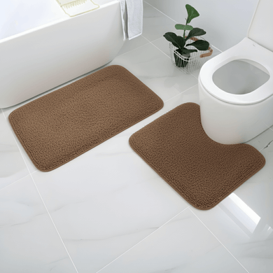 Brown bath mat set on a bathroom floor with a toilet and plant in the background.