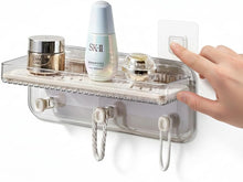 تحميل الصورة في عارض المعرض، A clear acrylic wall-mounted bathroom organizer shelf with white hooks, shown holding skincare bottles and jars, with a hand pressing a release button on the right side to detach it from a wall.

