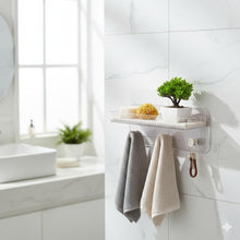 تحميل الصورة في عارض المعرض، A clear acrylic wall-mounted bathroom organizer with a small shelf and hanging hooks, shown attached to a white tiled wall in a bright bathroom. The organizer holds a small potted green plant, a yellow sponge, a bar of soap, and two towels hanging below.
