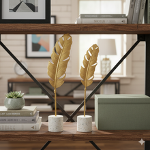 تحميل الصورة في عارض المعرض، Decorative gold feathers on white pedestals on a wooden shelf with books and a lamp in the background.
