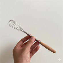 Load image into Gallery viewer, Hand holding a small whisk with a wooden handle against a plain background
