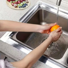 Load image into Gallery viewer, Person washing an orange under running water in a kitchen sink.
