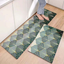 تحميل الصورة في عارض المعرض، Two rectangular anti-slip mats with a geometric leaf pattern in green and yellow shades, placed on a wooden floor. One mat is positioned in a kitchen setting with a person's feet in a relaxed pose on it.
