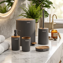 تحميل الصورة في عارض المعرض، A coordinated bathroom accessory set arranged on a marble countertop in a real bathroom setting, including a ribbed soap dispenser, cup, small container, lid, and toilet brush holder. The items are dark gray with light wood tops and small white SkyMaid branding.
