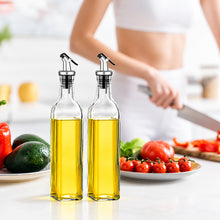 Load image into Gallery viewer, A clear glass bottle of olive oil with a black and silver pour spout, standing on a white countertop in a bright kitchen. A blurred person in white clothing is visible in the background, with plates of fresh vegetables nearby.
