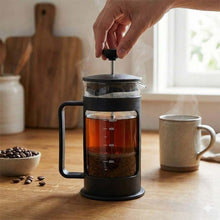 تحميل الصورة في عارض المعرض، Person using a French press to make coffee on a wooden table with a cup and coffee beans.
