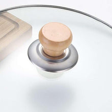 Load image into Gallery viewer, A close-up of a glass cookware lid featuring a round light wood knob and a brushed silver metal rim, shown on a clean white background.

