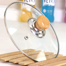 Load image into Gallery viewer, Clear glass pot lid with a light wood knob and stainless steel rim, resting on a wooden slatted stand.
