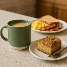 Load image into Gallery viewer, Green mug with coffee, plate of breakfast food including cake, eggs, and bacon on a countertop.
