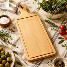 تحميل الصورة في عارض المعرض، A rectangular light wood cutting board with a handle and a round hole at the top, styled on a beige linen cloth with herbs, olives, salt, and pepper nearby.
