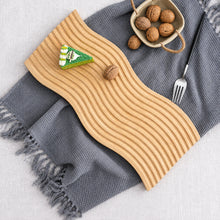 تحميل الصورة في عارض المعرض، A wavy, ribbed light wood serving board styled on a gray textured throw blanket with fringed edges. The board is decorated with a small green and white paper boat and a walnut, and is surrounded by a small woven basket of walnuts, a fork, and the gray blanket.
