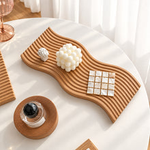 تحميل الصورة في عارض المعرض، A wavy wooden tray with concentric grooves and small square inlays, styled on a white round table with a white bubble candle, a small wrapped item, and a glass bottle nearby.
