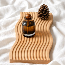 تحميل الصورة في عارض المعرض، Wooden tray with wavy design holding a glass bottle and pine cone on a white fabric background
