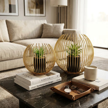 Load image into Gallery viewer, Two decorative gold wire globe planters, each holding a small green succulent, shown in two different settings: one on a reflective dark surface and one on a white surface against a light gray wall.
