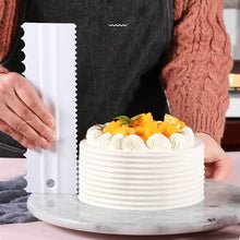 Load image into Gallery viewer, Person decorating a cake with a flexible icing scraper on a marble surface in a cozy kitchen setting.
