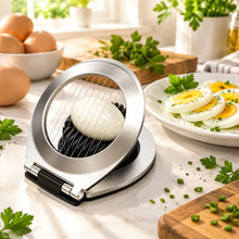 تحميل الصورة في عارض المعرض، A stainless steel egg slicer with a black handle, shown slicing a hard-boiled egg. A peeled egg and a plate of sliced hard-boiled eggs are visible in the background on a white studio background.
