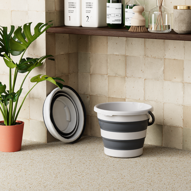 Collapsible gray and white bucket on a tiled floor with a plant and shelves in the background.