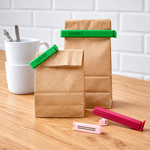Load image into Gallery viewer, Brown paper bags with green and pink clips on a wooden surface with white cups in the background.
