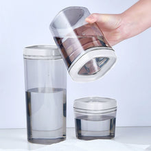 تحميل الصورة في عارض المعرض، Three translucent gray plastic water containers with white lids are shown on a light gray background, including one being held by a hand to demonstrate its tilt-up lid.
