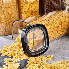 تحميل الصورة في عارض المعرض، A clear plastic food storage container with a black square lid and transparent window, shown tipped over with pasta spilling out onto a gray surface. Other pasta and grain containers are blurred in the background.
