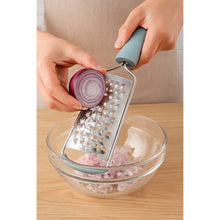 Load image into Gallery viewer, Person using a handheld grater to shred an onion over a glass bowl on a wooden surface.
