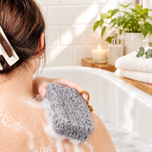 Load image into Gallery viewer, A woman shown from behind holding a pink textured sponge on her shoulder, with text reading 'Gentle decontamination' and 'Bright and radiant skin' over a soft white curtain background.
