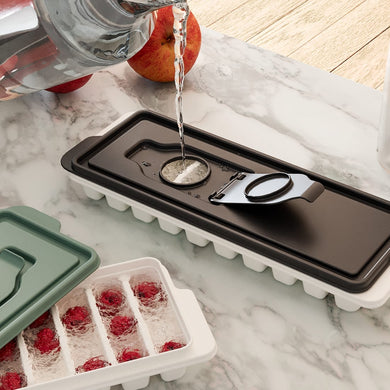 An ice cube tray with a black lid being filled with water, placed on a marble surface with apples in the background.