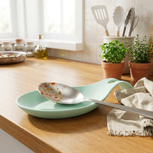 Load image into Gallery viewer, A mint green silicone spoon rest on a light wood kitchen counter, holding a stainless steel spoon with food residue, with kitchen utensils in the background.
