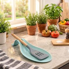 Load image into Gallery viewer, A silicone spatula with a wooden handle rests on a light blue spoon-shaped holder on a kitchen countertop, with a stovetop pot and kitchen items in the background.
