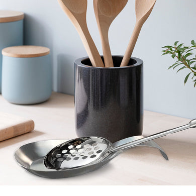 Black ceramic utensil holder with wooden spoons on a light surface, with a plant in the background.