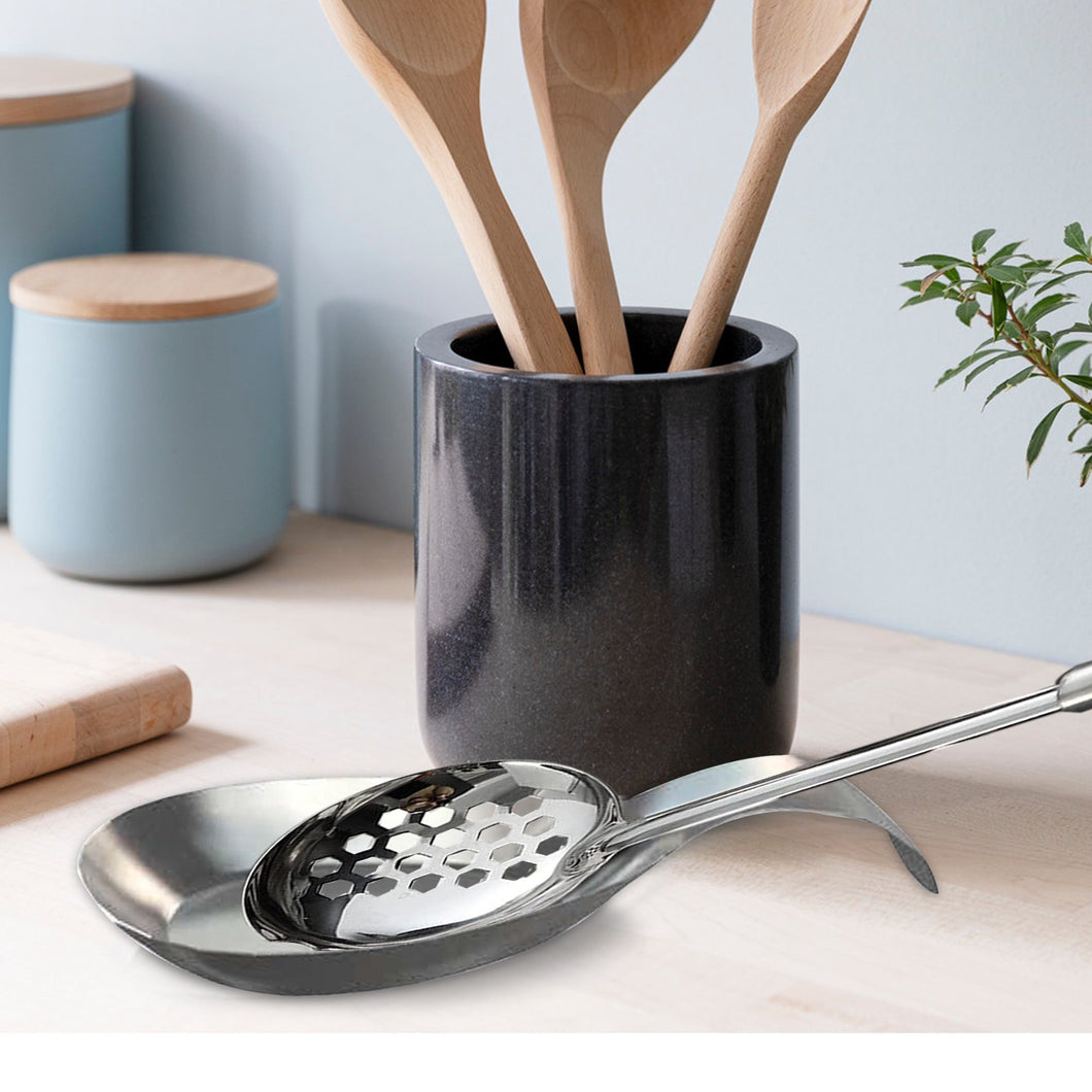 Black ceramic utensil holder with wooden spoons on a light surface, with a plant in the background.