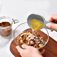 تحميل الصورة في عارض المعرض، Person pouring a liquid from a gray measuring spoon into a glass bowl with a brown substance on a white surface.
