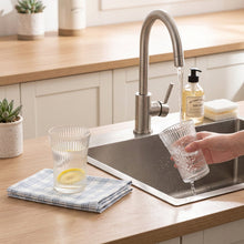 Load image into Gallery viewer, A three-panel collage showing a blue and white plaid reusable cleaning cloth being used in a kitchen sink to wash a clear ribbed glass cup, with the cloth also placed on a tabletop next to the cup.
