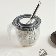 Load image into Gallery viewer, A gray mixing bowl with a lid and measurement lines, filled with a white substance, possibly flour or sugar, and a metal whisk inside the bowl. The bowl is placed on a table with a cup and a spoon beside it.

