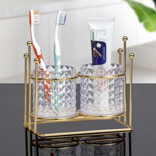 تحميل الصورة في عارض المعرض، Gold toothbrush holder with two cups, toothbrushes, and a tube of toothpaste on a gray surface.
