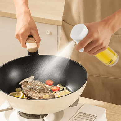 A person is using a clear glass oil sprayer with a white pump to spray oil over food in a pan.
