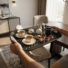 تحميل الصورة في عارض المعرض، A person holds a black perforated serving tray in a bright dining room. The tray is loaded with two teacups on saucers, a small teapot, a cream-colored teapot, and several small bowls of snacks and sweets.
