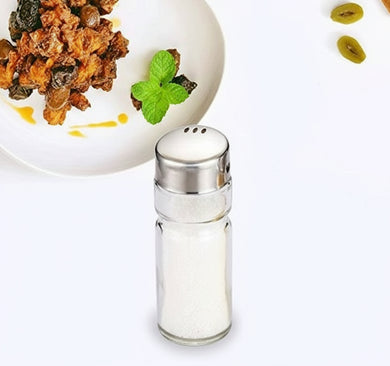 Two glass salt and pepper shakers on a white background with a dish of food in the background.