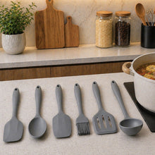 Load image into Gallery viewer, A set of seven gray silicone kitchen utensils, including spatulas, a brush, and a ladle, arranged on a light beige countertop next to a pot of soup on an induction cooktop. The background shows a styled kitchen with wooden cutting boards, jars, and plants.
