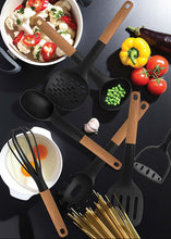 تحميل الصورة في عارض المعرض، A set of black silicone kitchen utensils with light brown wooden handles, arranged on a dark surface with various food ingredients like tomatoes, peas, garlic, pasta, and a pot of pasta.
