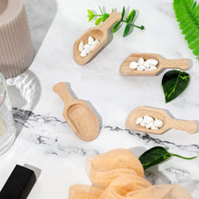 Load image into Gallery viewer, Four small natural wood scoop-like containers arranged on a white marble surface with decorative leaves, a beige candle, a glass, and a peach mesh loofah.
