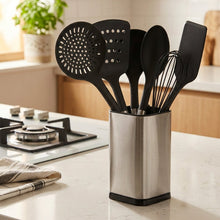 Load image into Gallery viewer, A hand holding a stainless steel kitchen utensil holder filled with black silicone kitchen tools, including a spatula, spoon, and whisk, in front of a store shelf background.
