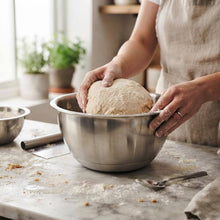 Load image into Gallery viewer, A person wearing a beige apron kneads a ball of dough inside a large stainless steel mixing bowl on a marble kitchen counter.
