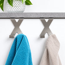 Load image into Gallery viewer, Two stainless steel X-shaped towel hooks mounted on a white wall beneath a gray shelf, with a turquoise towel hanging on the left and a light beige towel hanging on the right.
