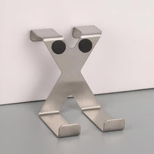 Load image into Gallery viewer, A brushed stainless steel X-shaped wall hook with two circular black magnets on the upper crossbar, shown standing on a gray surface against a white wall.
