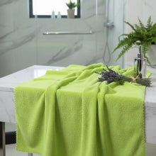 Load image into Gallery viewer, Green towel draped over a bathtub with a plant and toiletries in the background
