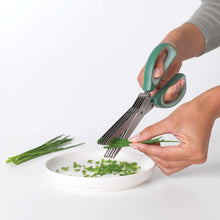 Load image into Gallery viewer, Person using kitchen shears to chop green herbs on a white plate with a white background
