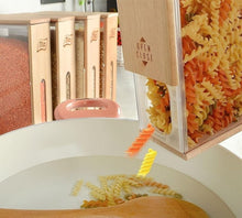 Load image into Gallery viewer, A wooden pasta maker device with clear panels showing rotini pasta being poured into a white bowl, with spice containers blurred in the background.
