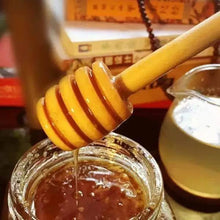 تحميل الصورة في عارض المعرض، A wooden honey dipper dripping thick golden honey into a glass jar, with a small pitcher and blurred books in the background.
